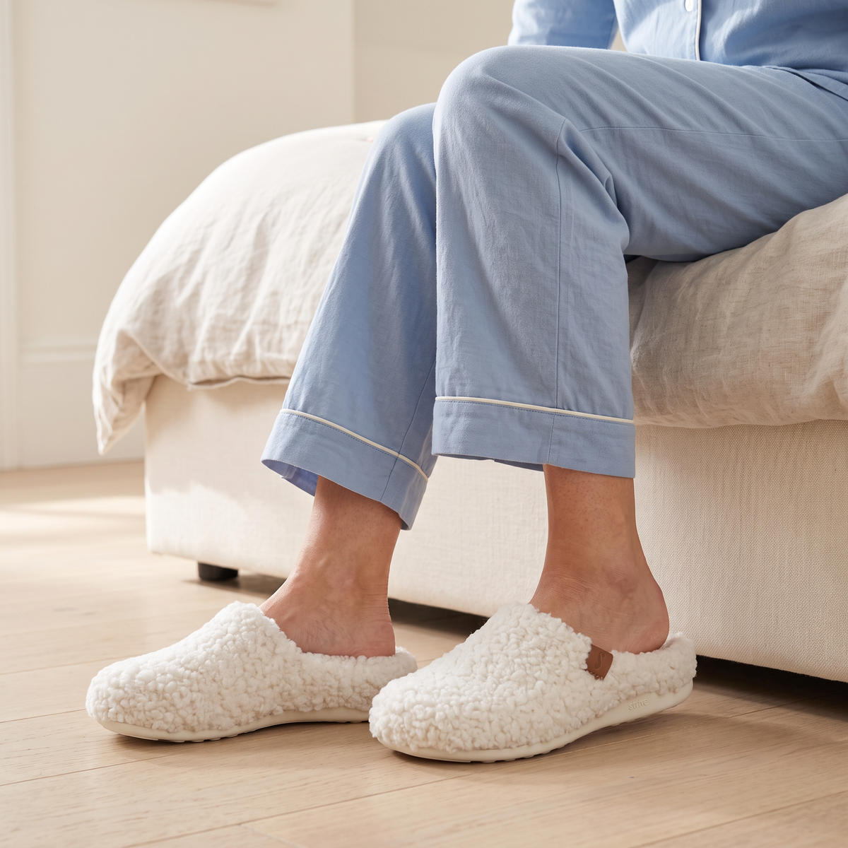 Woman in blue pajamas sitting on the edge of a bed wearing Strive Copenhagen II Curly Marshmallow white orthotic slippers.